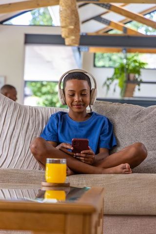Adolescent boy using smartphone while wearing headphones on cozy living room couch