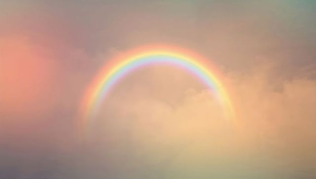 Glowing pastel rainbow arching across soft peach and pink cloudscape at golden hour