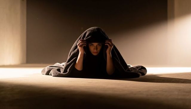 Hooded woman hiding under blanket on concrete floor in dim industrial light, solitude