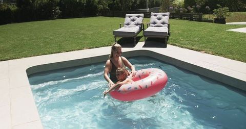 Parent supporting child on pink donut float playing in sunlit backyard swimming pool