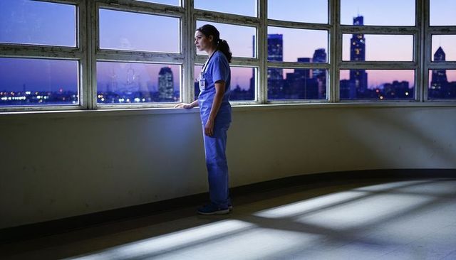 Night-shift nurse in blue scrubs standing at window gazing at city skyline at dusk