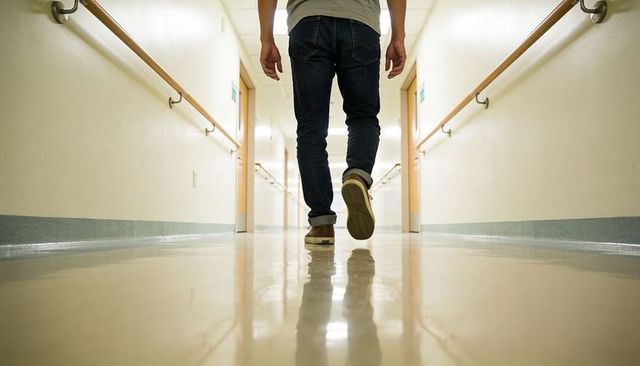 Man walking down hospital corridor with reflective floor and handrails, casual footwear