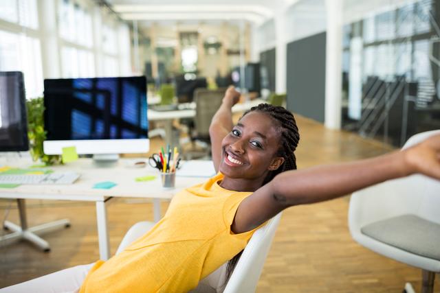 Young female graphic designer stretching and smiling at her office desk. Ideal for use in articles or advertisements about workplace happiness, creative professions, modern office environments, and productivity. Can also be used for promoting ergonomic office furniture and employee wellness programs.