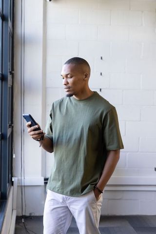 African american man in his 20s standing by window checking smartphone in modern loft