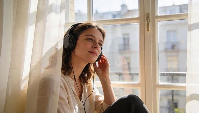 Woman listening with headphones at window seat gazing through sheer curtains in daylight