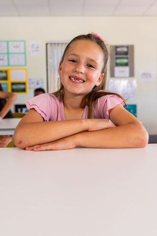 Smiling schoolgirl showing missing tooth at classroom desk wearing pink top
