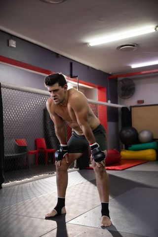 Shirtless mma fighter leaning forward breathing heavily inside training cage