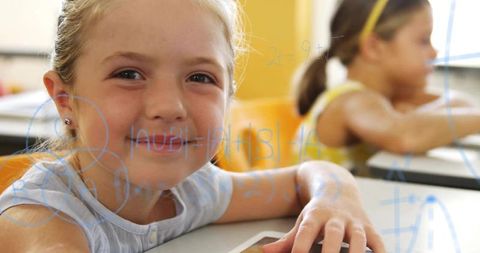 Smiling blonde girl using tablet in classroom with math overlay, digital learning concept