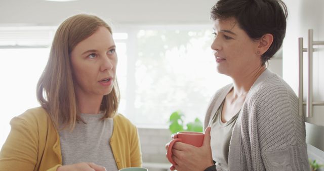 Two women are seen engaging in conversation while holding coffee mugs in a casual home kitchen environment. This image can be used to illustrate themes such as friendship, daily life, and social interaction. Suitable for articles on relationships, lifestyle blogs, or content aimed at portraying social dynamics and home settings.