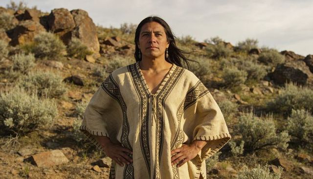 Standing indigenous man on sunlit desert hillside wearing woven poncho and earrings