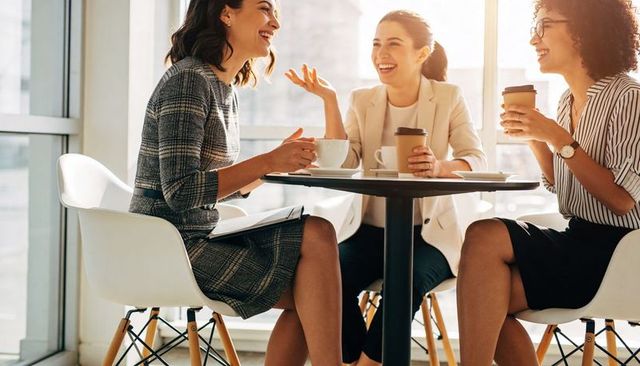 Three professional women laughing and networking over coffee in modern office café