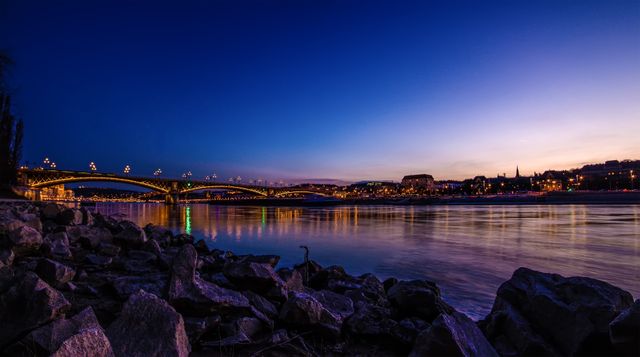 Captures the mesmerizing twilight over a calm river with a bridge illuminated by night lights, reflecting in the water. Ideal for use in travel blogs, tourism promotions, cityscapes brochures, or websites focusing on scenic landscapes and urban beauty.