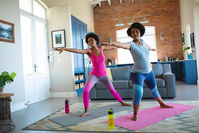 African American mother and daughter practicing yoga together in a modern living room. Both are wearing colorful workout clothes and are positioned on yoga mats, with water bottles nearby. Ideal for promoting family fitness, home workouts, healthy living, and mother-daughter bonding activities.
