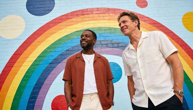Two men standing before vibrant rainbow mural wearing casual summer button-up shirts