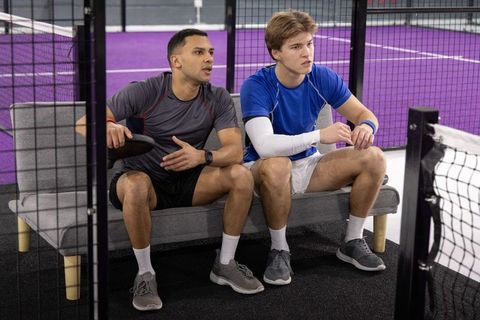 Male teammates sitting on bench holding paddles watching pickleball match on purple court