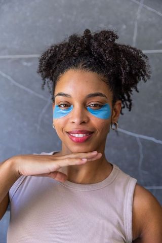 African american woman wearing blue under-eye patches, smiling and showcasing glowing skincare routi