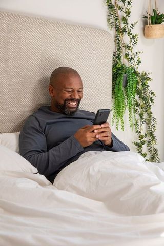 Mature african american man reclining in bed smiling and scrolling smartphone near hanging plants