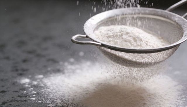 Sifting flour through fine mesh sieve over dark stone countertop creating powdered pile