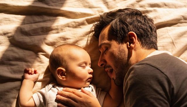 Father sleeping beside baby on sunlit bed, tender bonding moment and morning calm