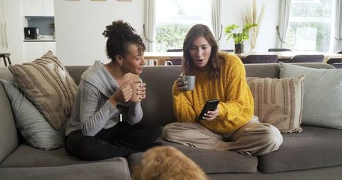 Diverse friends laughing and sharing smartphone while drinking coffee on cozy sofa
