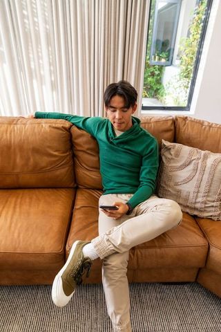 Asian man sitting on caramel leather couch using smartphone in cozy modern living room