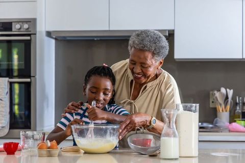 African grandmother teaching granddaughter baking while whisking batter in modern kitchen