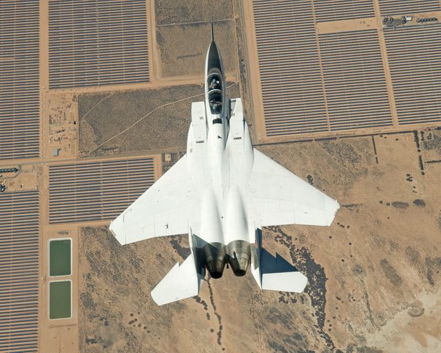 Top-down view of F-15D fighter jet flying inverted over Mojave Solar Project in Southern California desert. White aircraft contrasts with desert terrain and orderly grid of solar panels. Can be used in aerospace and defense content, showcasing military prowess, aviation technology, or renewable energy programs.