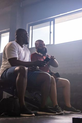 African men sitting on gym bench checking phones and smartwatch during workout rest