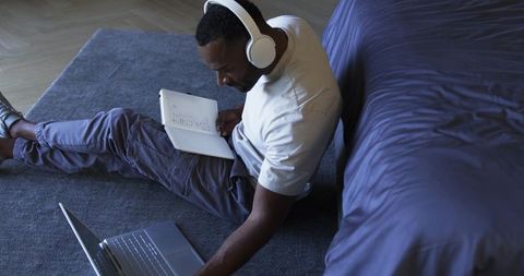 Focused man studying with headphones taking handwritten notes and using laptop on bedroom floor
