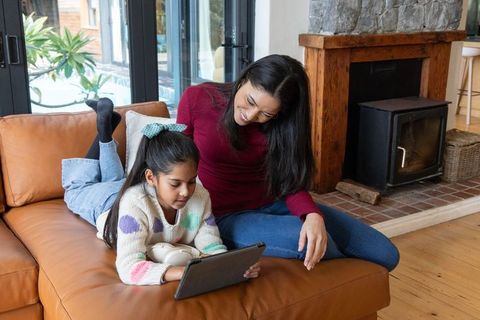 Indian mother and daughter sharing tablet on leather sofa by fireplace, cozy family moment