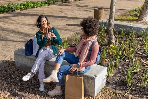 Friends sharing smartphone while sitting on concrete bench in sunny urban park
