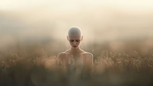 Shaved-head woman standing in dew-soaked meadow at golden hour, meditative solitude