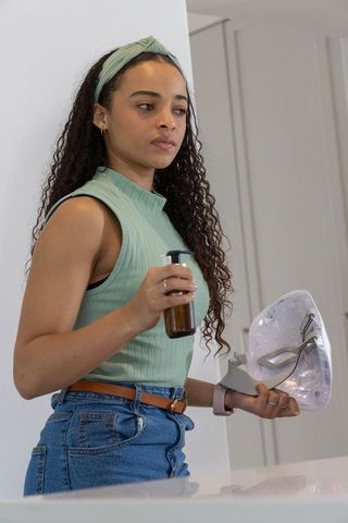 Young african woman holding amber serum pump and led facial mask wearing mint top and jeans
