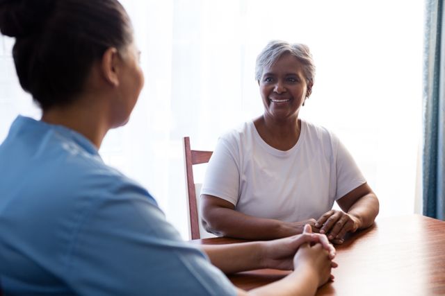 Nurse interacting with senior woman at table in nursing home. Ideal for use in healthcare, elderly care, and medical service promotions. Can be used in brochures, websites, and advertisements focusing on senior care, nursing homes, and healthcare services.