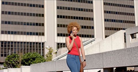 African American woman walking and talking on phone in sunlit downtown plaza casual style