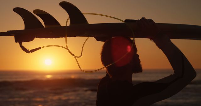 Surfer holding surfboard above head at sunset on beach. Silhouette against golden sky, capturing the serenity of surfing lifestyle. Ideal for promoting summer vacations, outdoor activities, water sports, travel destinations, or healthy living.