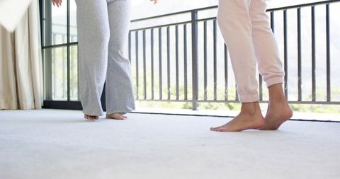 Sunlit diverse barefoot friends in loungewear standing on soft carpet near balcony rail