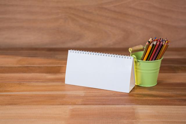 Blank spiral calendar standing on wooden table next to a green bucket filled with colored pencils. Ideal for use in office, school, or home settings to depict organization, planning, or creative activities. Perfect for advertisements, blog posts, or educational materials.