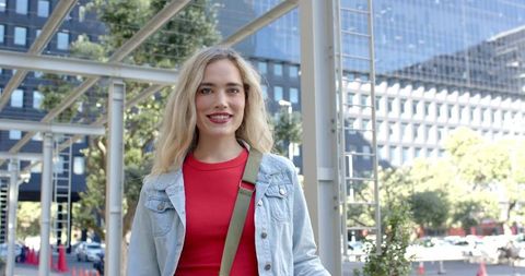 Young woman smiling on urban sidewalk wearing red top and denim jacket holding green bag