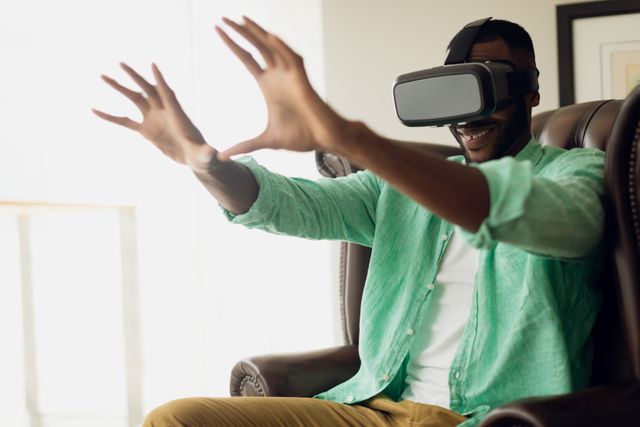 African-American man sitting on a leather chair in a room, wearing virtual reality goggles and reaching out with his hands. Ideal for use in technology blogs, virtual reality product promotions, gaming advertisements, and articles about modern home entertainment.