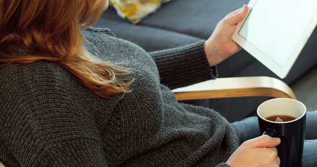 Woman sitting comfortably on couch using tablet while holding tea mug. Great for topics on relaxation, digital lifestyle, technology dependence, home comfort, and casual moments.