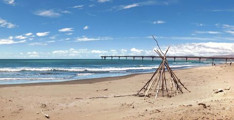 Driftwood Teepee on Sandy Beach with Long Pier Stretching into Calm Blue Ocean Horizon