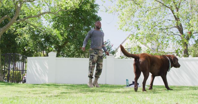 African american male soldier exercising and jumping rope with dog in garden. Patriotism, army and returning home concept.