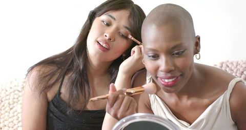 Friends applying makeup together on sofa sharing brushes and mirror for beauty routine