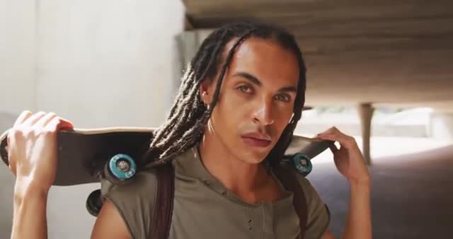 A young man with long dreadlocks confidently holds a skateboard over his shoulders while standing in a vibrant city environment. This image, with its energetic and stylish vibe, is ideal for use in fashion editorials, urban lifestyle promotions, or advertising targeting youthful, modern audiences. The underpass and clear ambiance offer dramatic angles that highlight urban living.