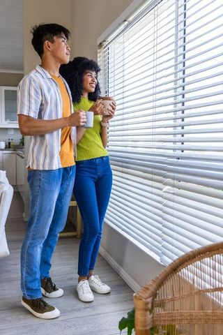 Young couple standing by window holding coffee mugs and looking out through blinds