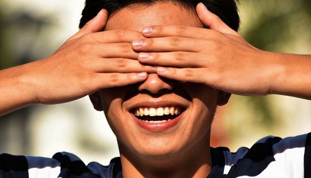 Teen smiling while covering eyes with hands in sunlit park casual striped shirt