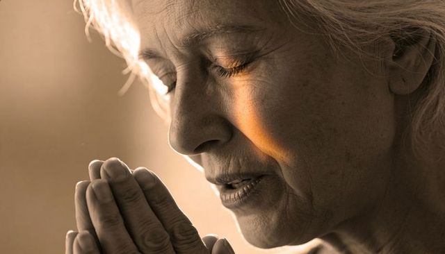Elderly asian woman praying with hands near mouth in warm sunlight, contemplative portrait