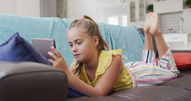 Young girl in a casual setting lies comfortably on a sofa, using a tablet to stay connected. Ideal for themes related to childhood, digital activities, leisure time, and everyday home life. Useful for educational content, technology use among children, and lifestyle blogs.