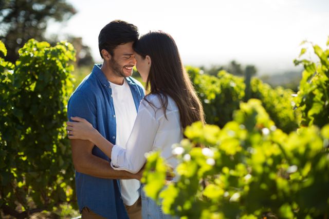 Young couple standing close and smiling in a lush vineyard on a sunny day. Ideal for use in advertisements, travel brochures, romantic getaway promotions, lifestyle blogs, and social media content focusing on love and relationships.